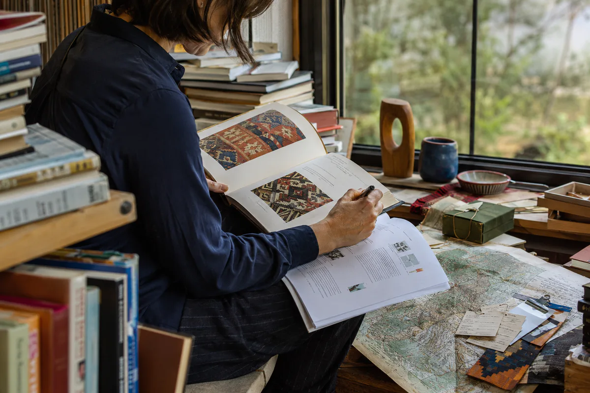 A researcher cross-checking an illustrated reference book and printed report among maps, notes and material samples.